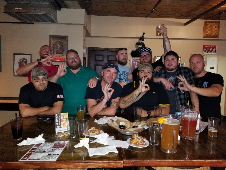A group of nine men poses for a photograph behind a table holding snacks and pitchers of beer. Some of the men are in Proud Boy shirts. Several of them are making the "ok" sign with their hands.